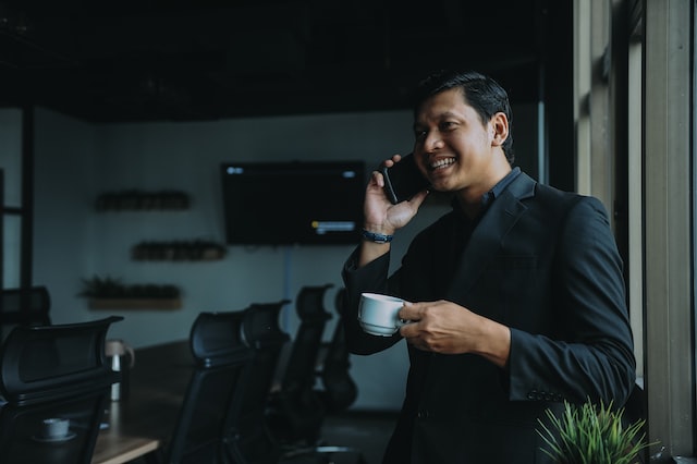 A man standing in an office talks on the phone while holding a cup of coffee. 
